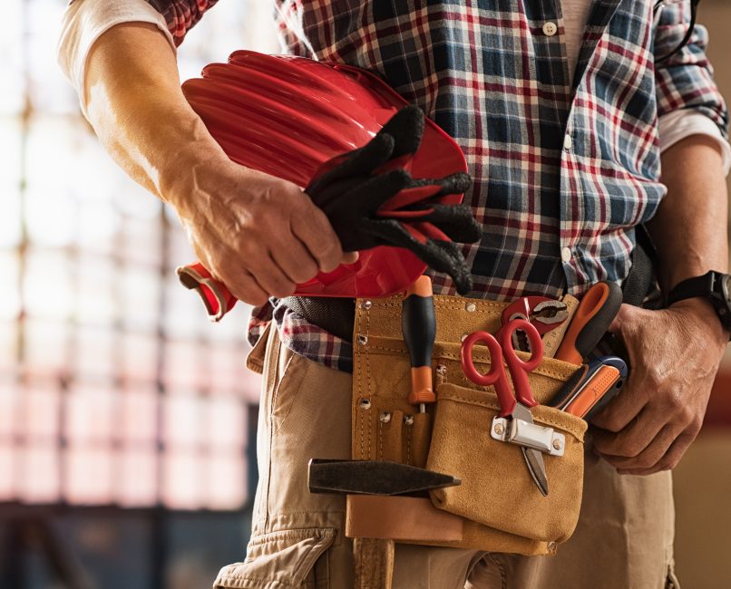 Closeup of bricklayer hands holding hardhat and construction equipment. Detail of mason man hands holding work gloves and wearing tool kit on waist. Handyman with tools belt and artisan equipment.